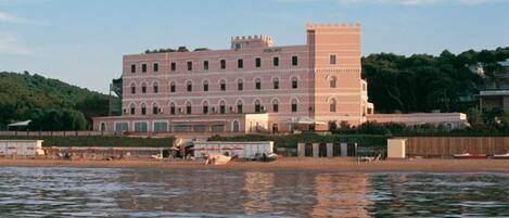 A large, historic building with multiple arches and a prominent tower, situated by a body of water.