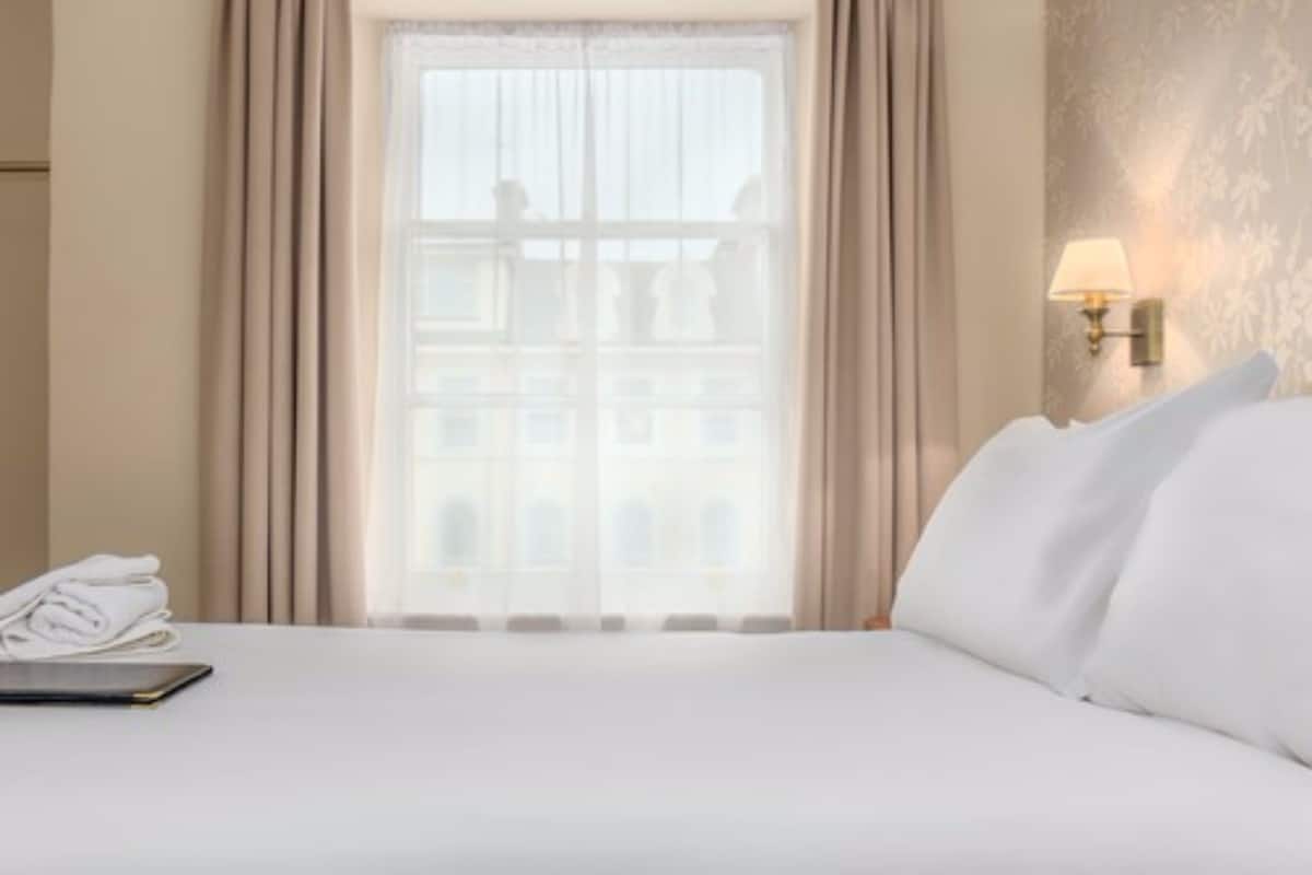 A hotel room with a neatly made bed, a window with curtains, a lamp on the wall, and a folded towel on the side table.