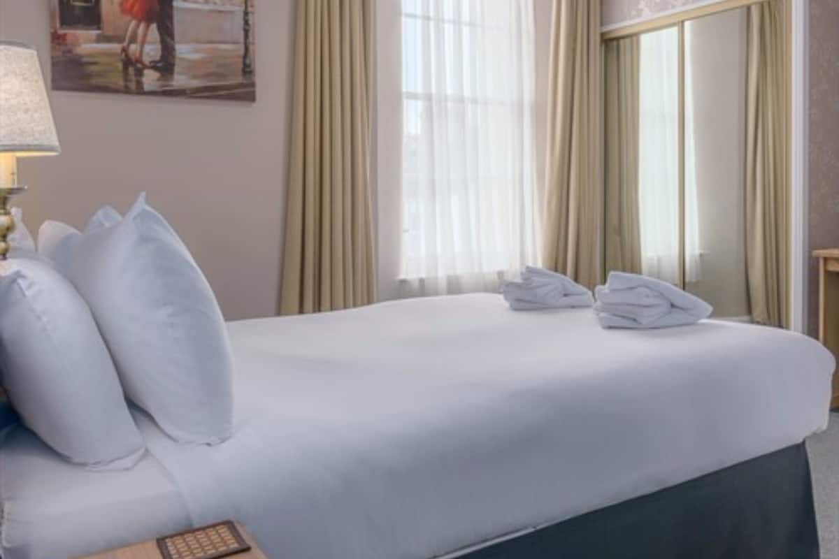 A hotel room with a bed, pillows, a folded towel, a remote control, and a painting on the wall.