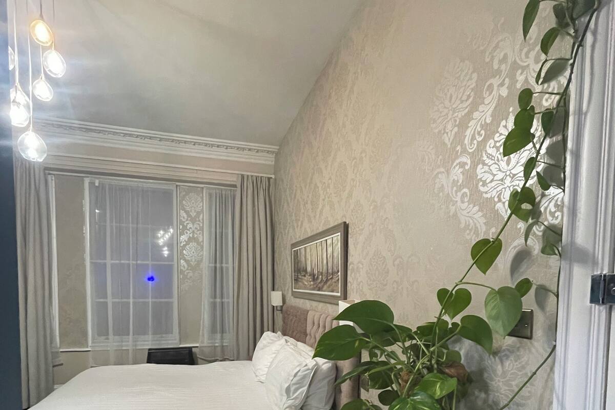 A bedroom with a bed, a potted plant, a nightstand, a window with curtains, and a wall with a framed picture.