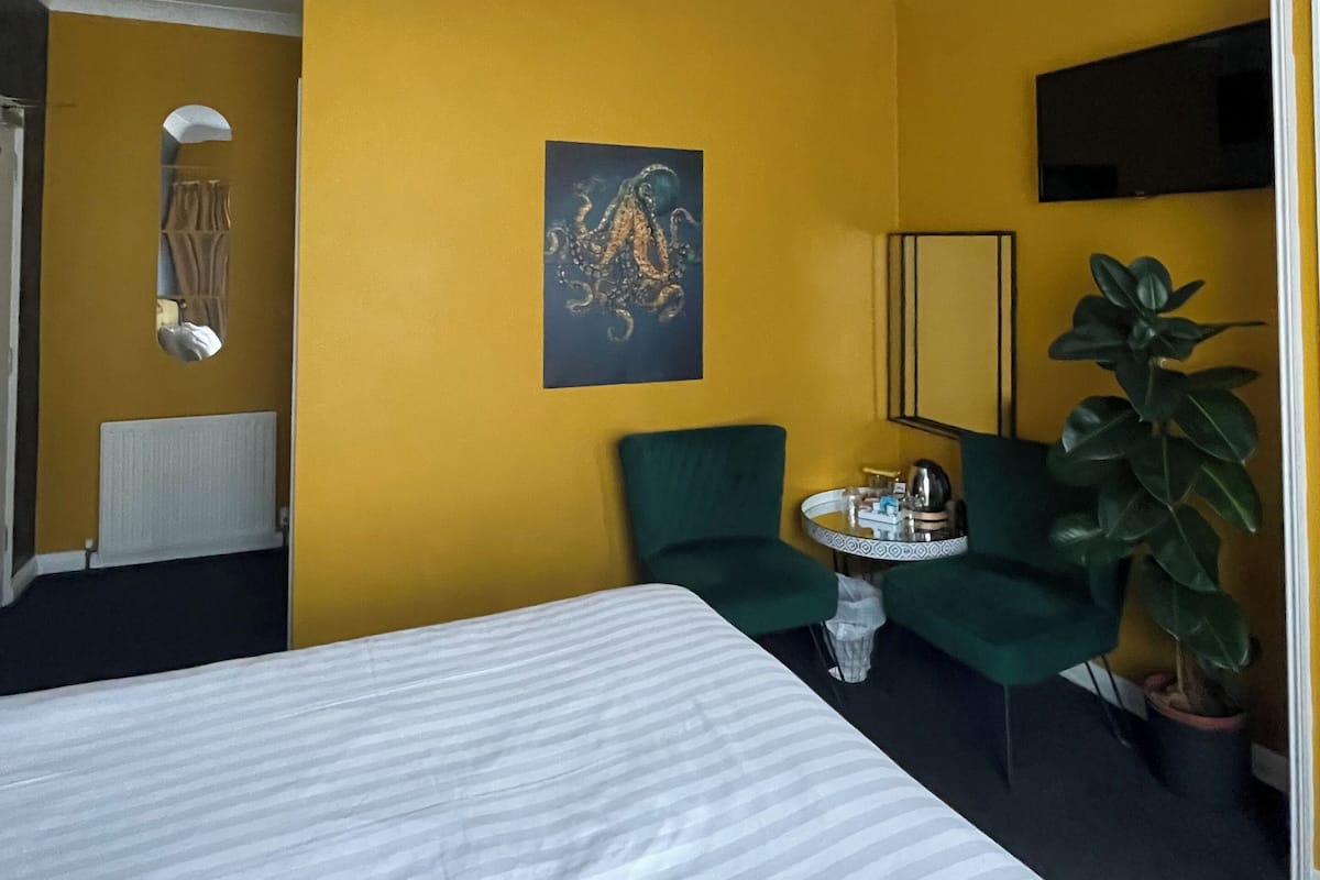 A hotel room with a bed, a ceiling fan, a painting, a green chair, a small table with a tray, and a potted plant.