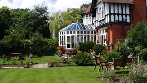 A traditional Tudor-style building with a conservatory, surrounded by a well-maintained garden with various plants and a lawn. - The Normanhurst Hotel (Sale)
