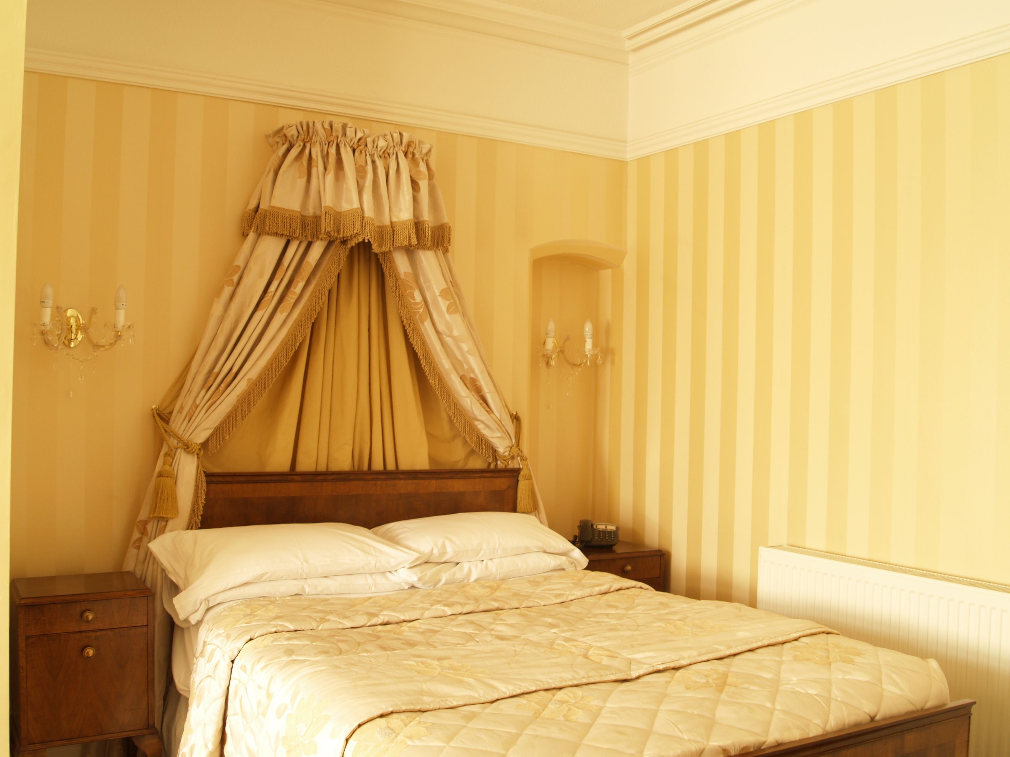 A bedroom with a canopy bed, a nightstand, a bedside table, and a wall-mounted lamp.