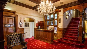 A reception area with a wooden desk, a leather chair, a chandelier, and a red carpeted staircase. - The Normanhurst Hotel (Sale)
