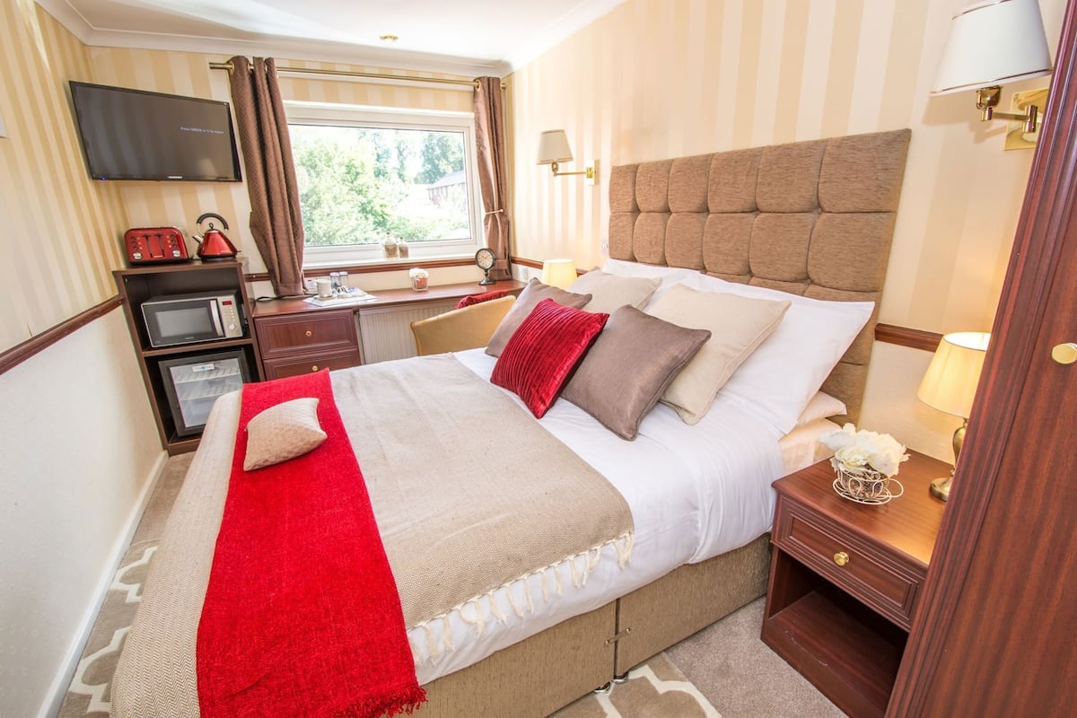 A hotel room with a bed, a TV, a desk with a kettle, a lamp, and a window with curtains.