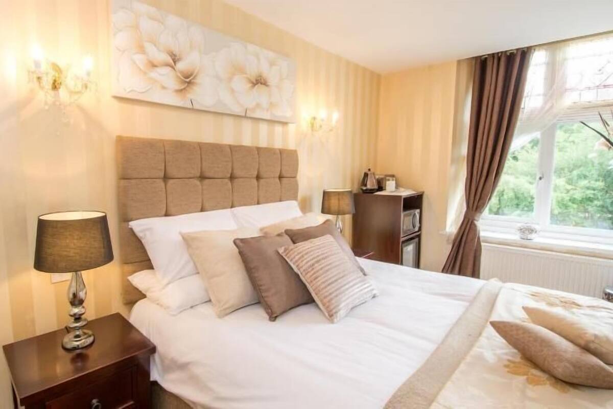 A hotel room with a large bed, a nightstand with a lamp, a wall-mounted artwork, and a window with curtains.