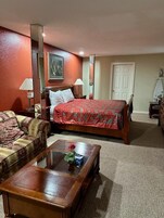 Habitación de hotel con una cama, una mesa de centro de madera con un jarrón de flores, un sofá y un cuadro en la pared.