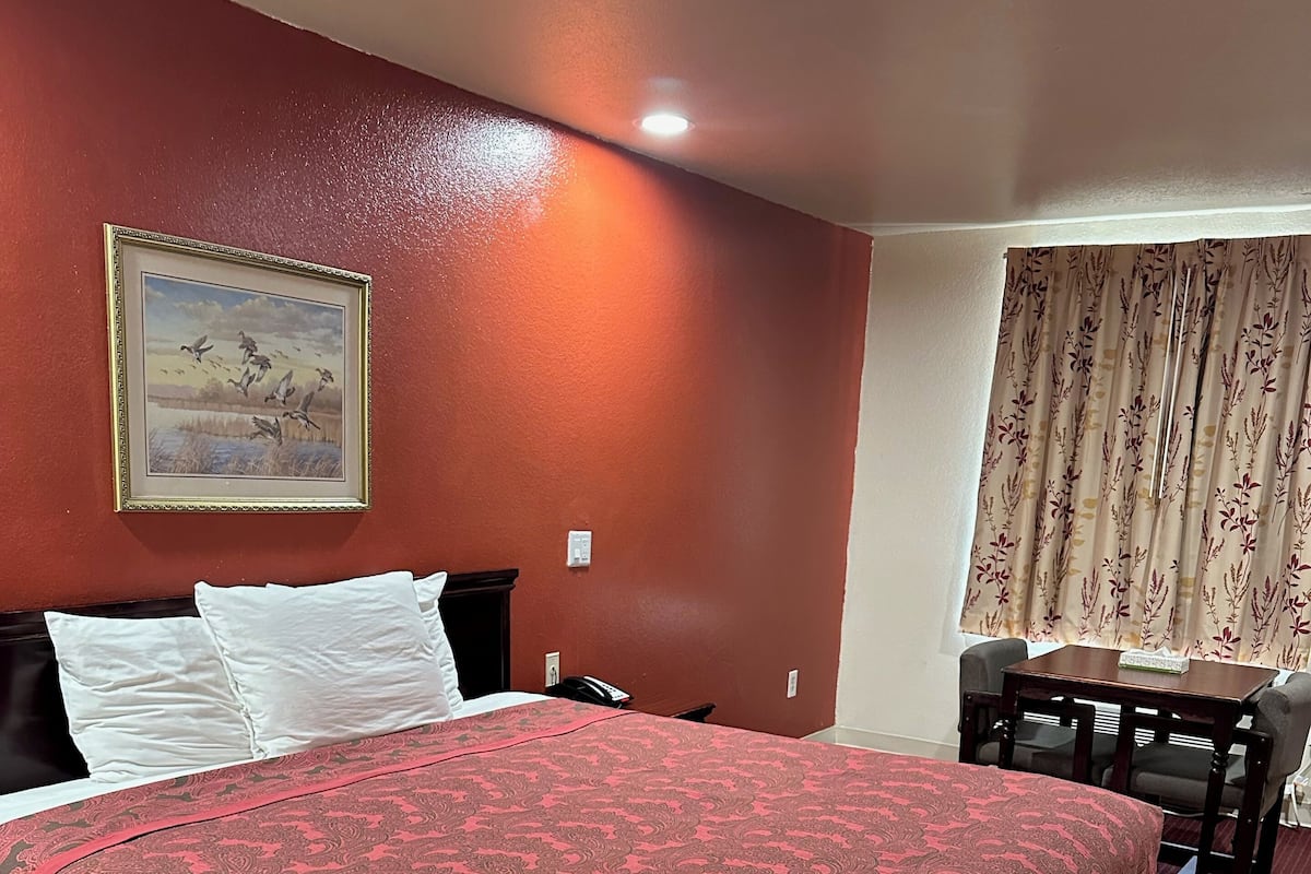 Une chambre d’hôtel avec un grand lit, un bureau et une chaise. Il y a un tableau au mur et une fenêtre avec des rideaux.