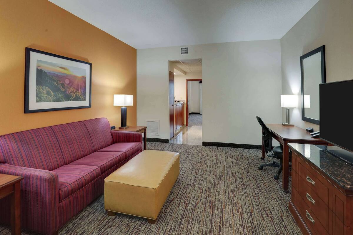 Een hotelkamer met een gestreepte bank, een klein poufje, een bureau met een computer, een televisie en een lijst aan de muur.