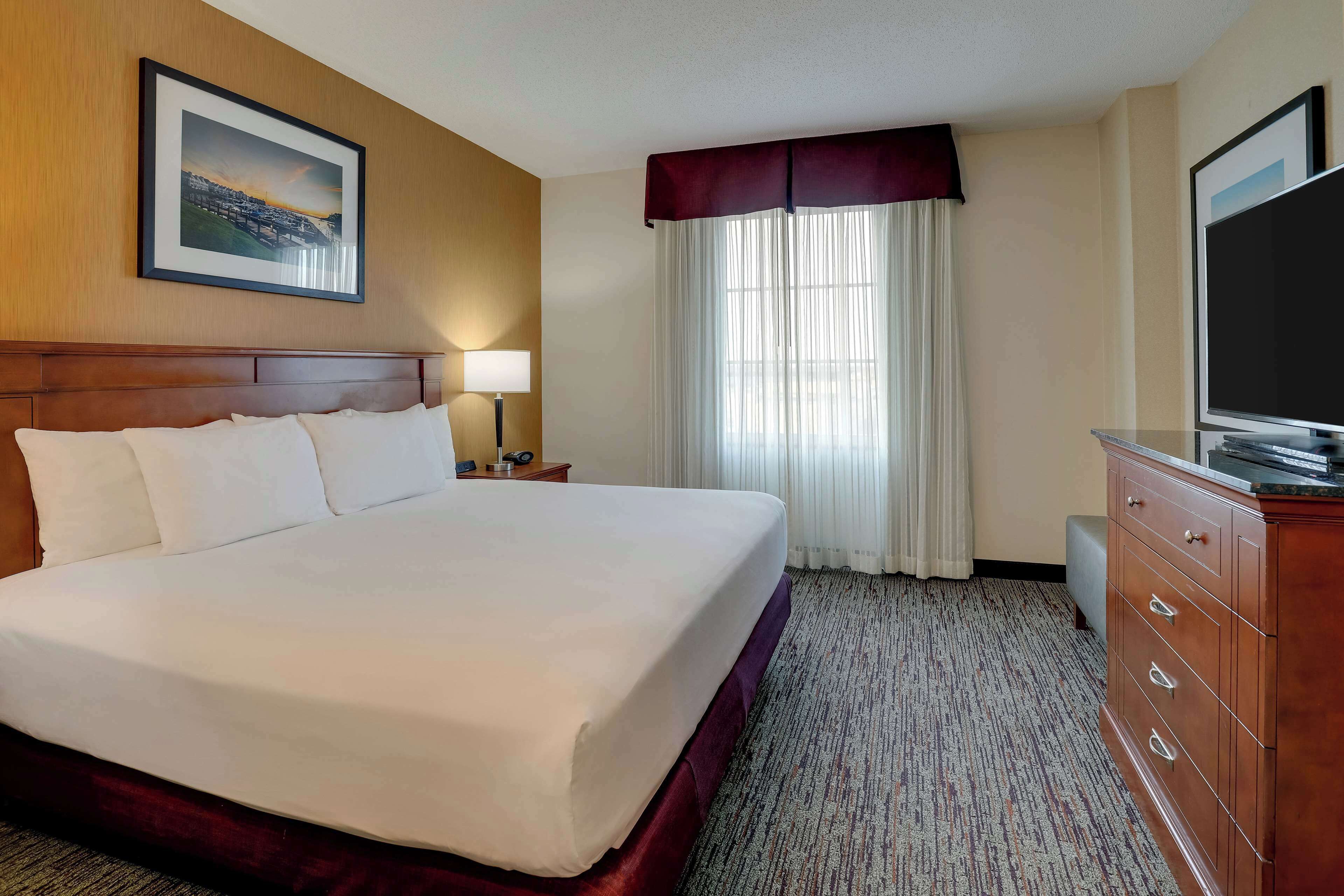 A hotel room with a large bed, a wooden headboard, a flat-screen TV, a dresser, a nightstand with a lamp, and a framed picture on the wall.