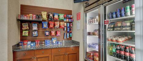 A snack bar with a variety of packaged snacks and beverages, a television mounted above, and a refrigerator stocked with drinks.