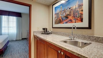 A hotel room with a kitchenette, a bed, a window with curtains, and a cityscape painting on the wall.