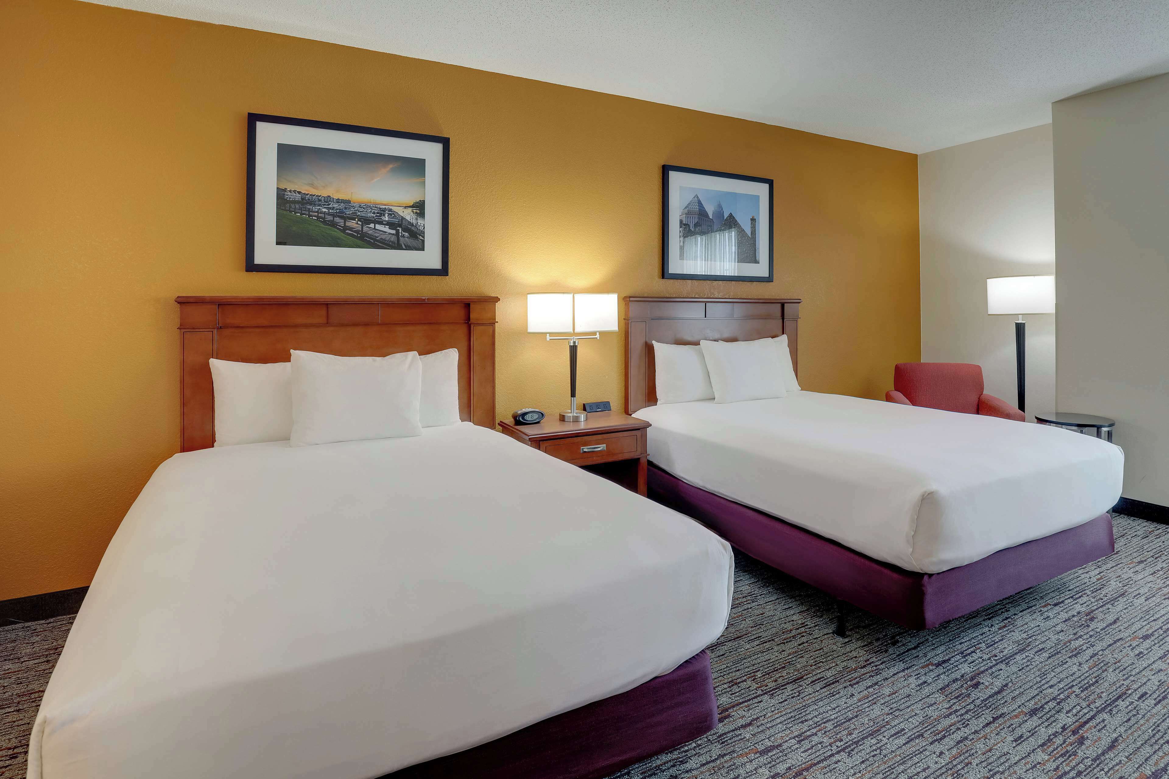 A hotel room with two beds, a wooden headboard, a nightstand with a lamp, and two framed pictures on the wall.