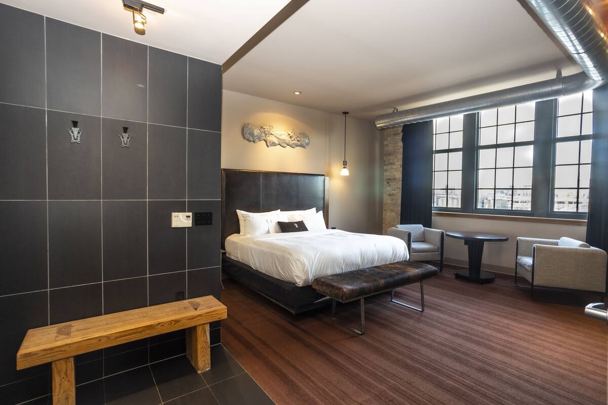 Ein modernes Hotelzimmer mit einem großen Bett, einer Bank, einem kleinen Tisch und einer Sitzecke am Fenster.