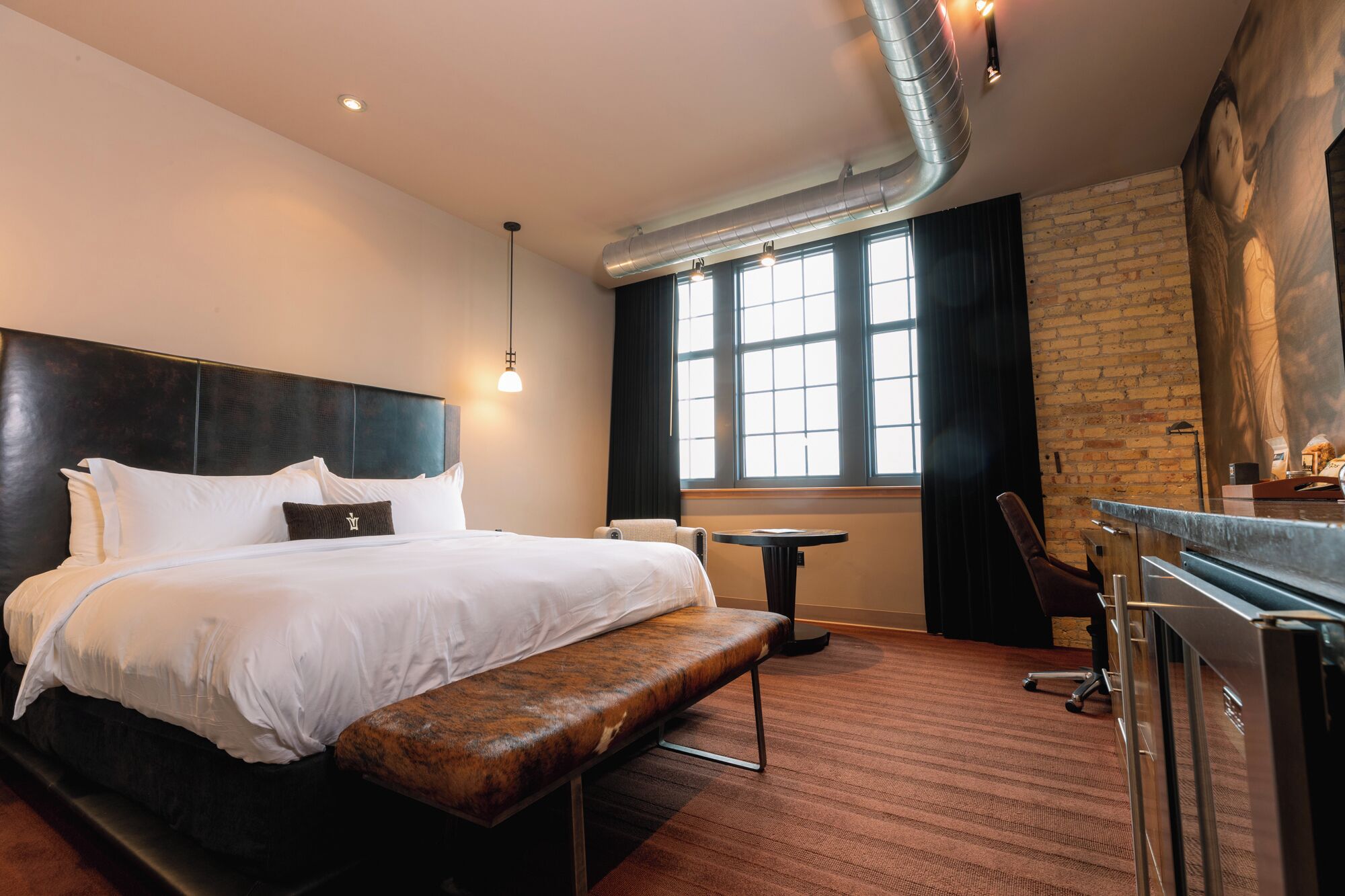 A hotel room with a large bed, a bench, a small table, a desk, and a large window.