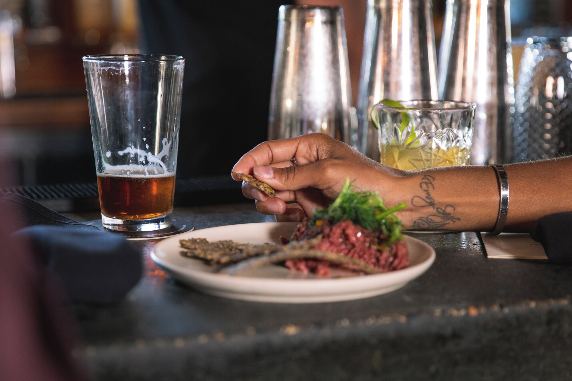 A hand holding a cracker next to a plate with a meat dish and a glass of beer.