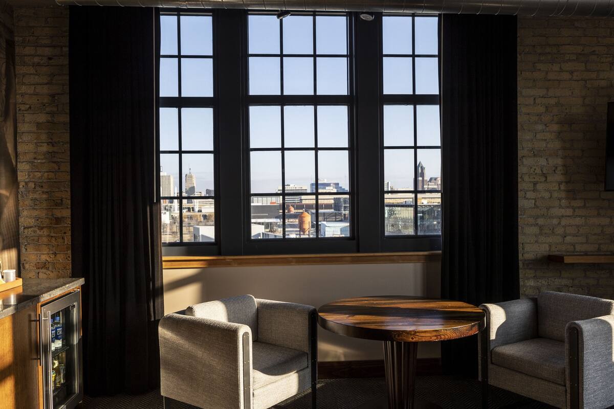 Ein Zimmer mit großem Fenster, zwei Sesseln, einem kleinen runden Tisch und einer Minibar.