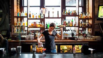 A bartender preparing a drink at a bar with shelves of liquor bottles in the background.