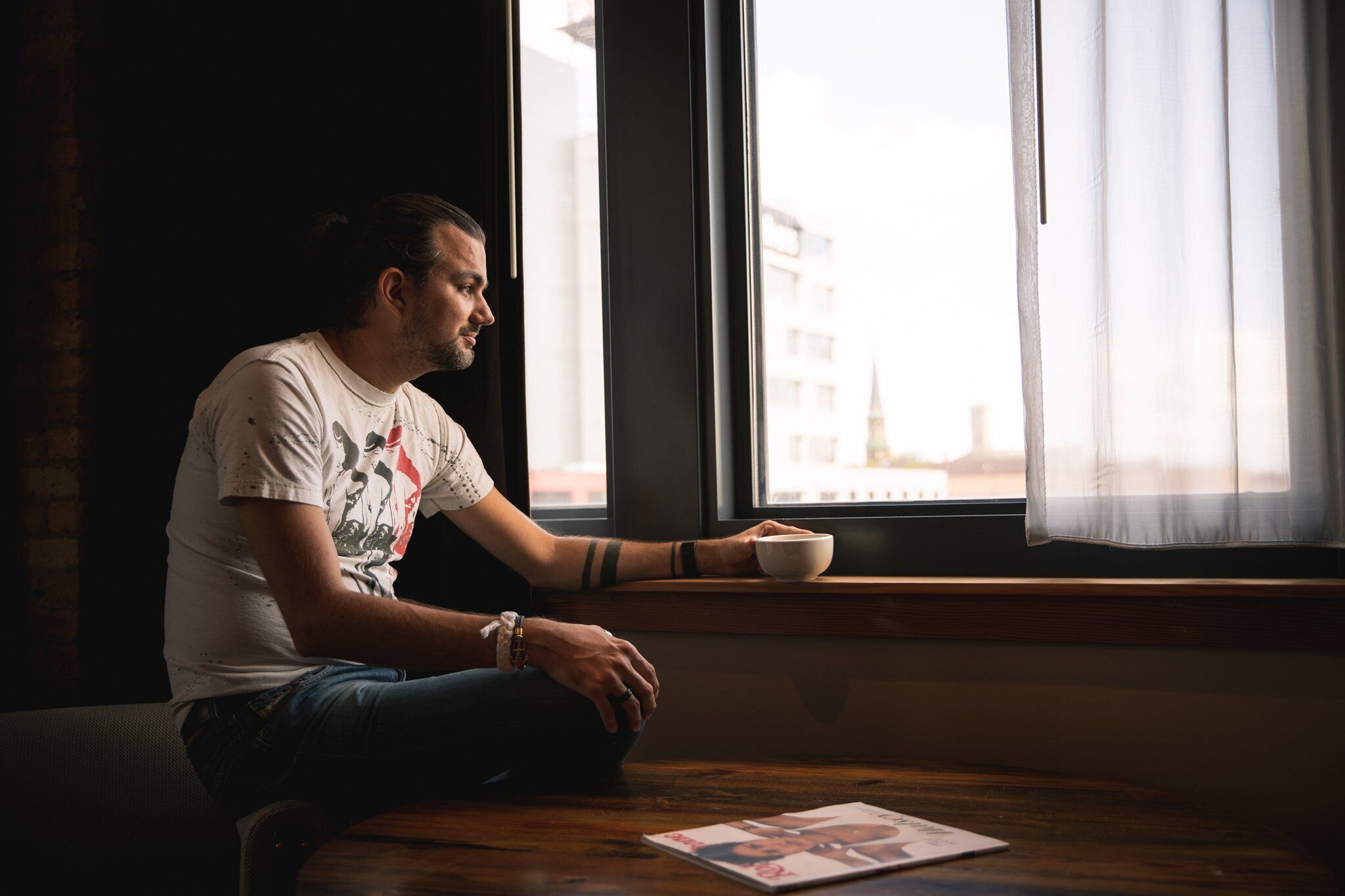 A person sitting by a window with a bowl, a watch, and a magazine on a table.