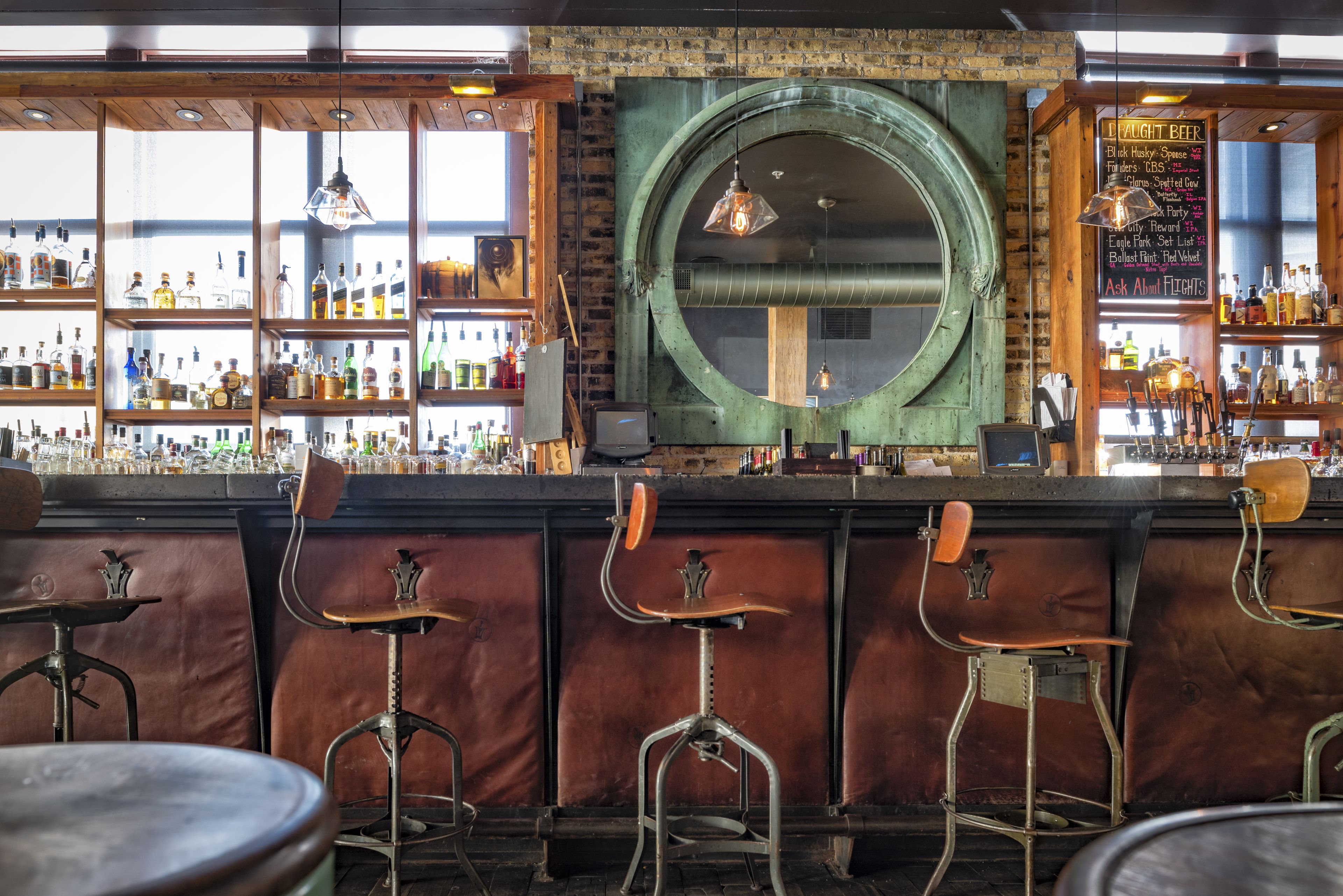 A bar with stools, shelves of bottles, and a large circular window.