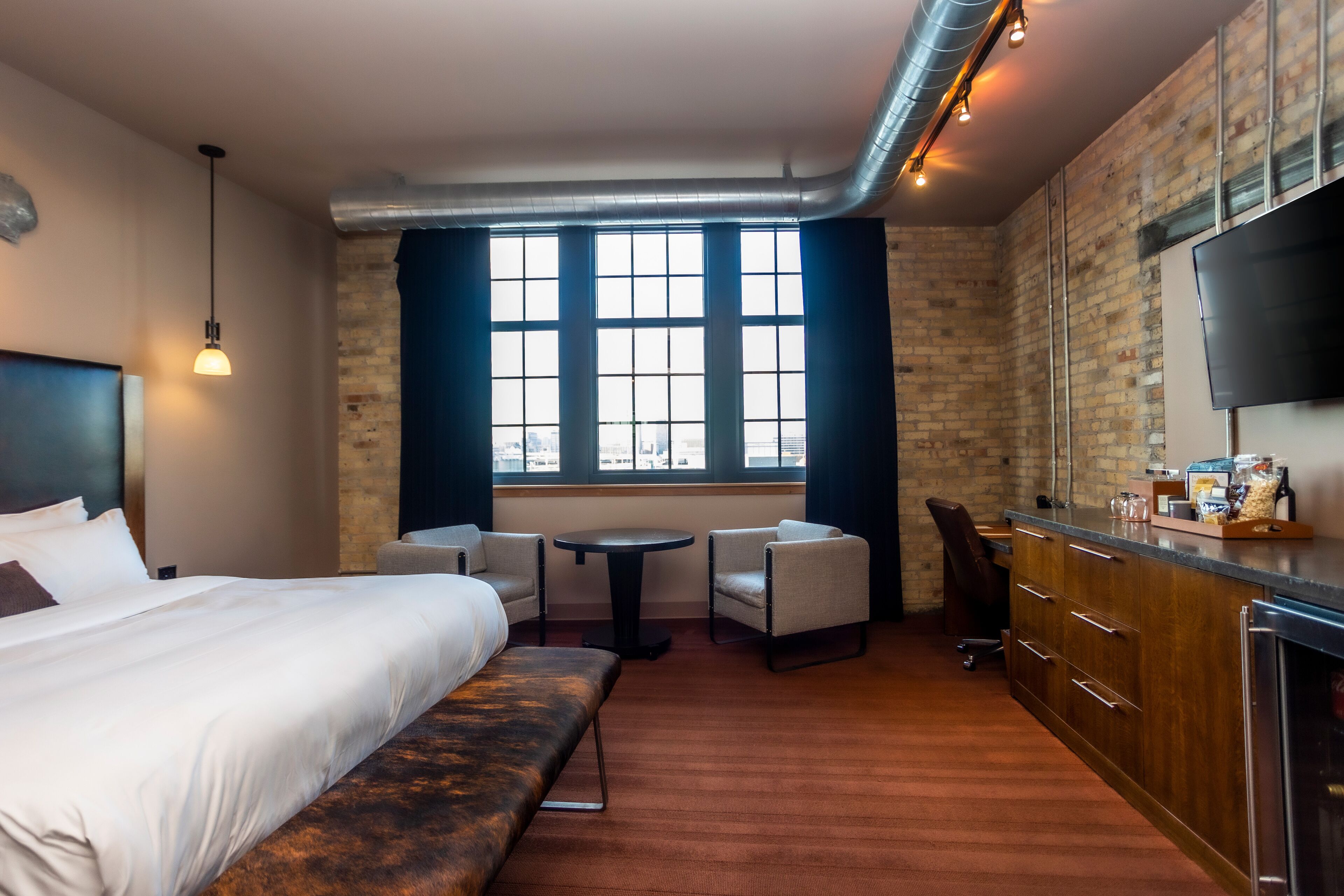 Habitación de hotel con una cama grande, una banca, una mesita redonda pequeña, dos sillones, un televisor y un área de cocina con mostrador y armarios.