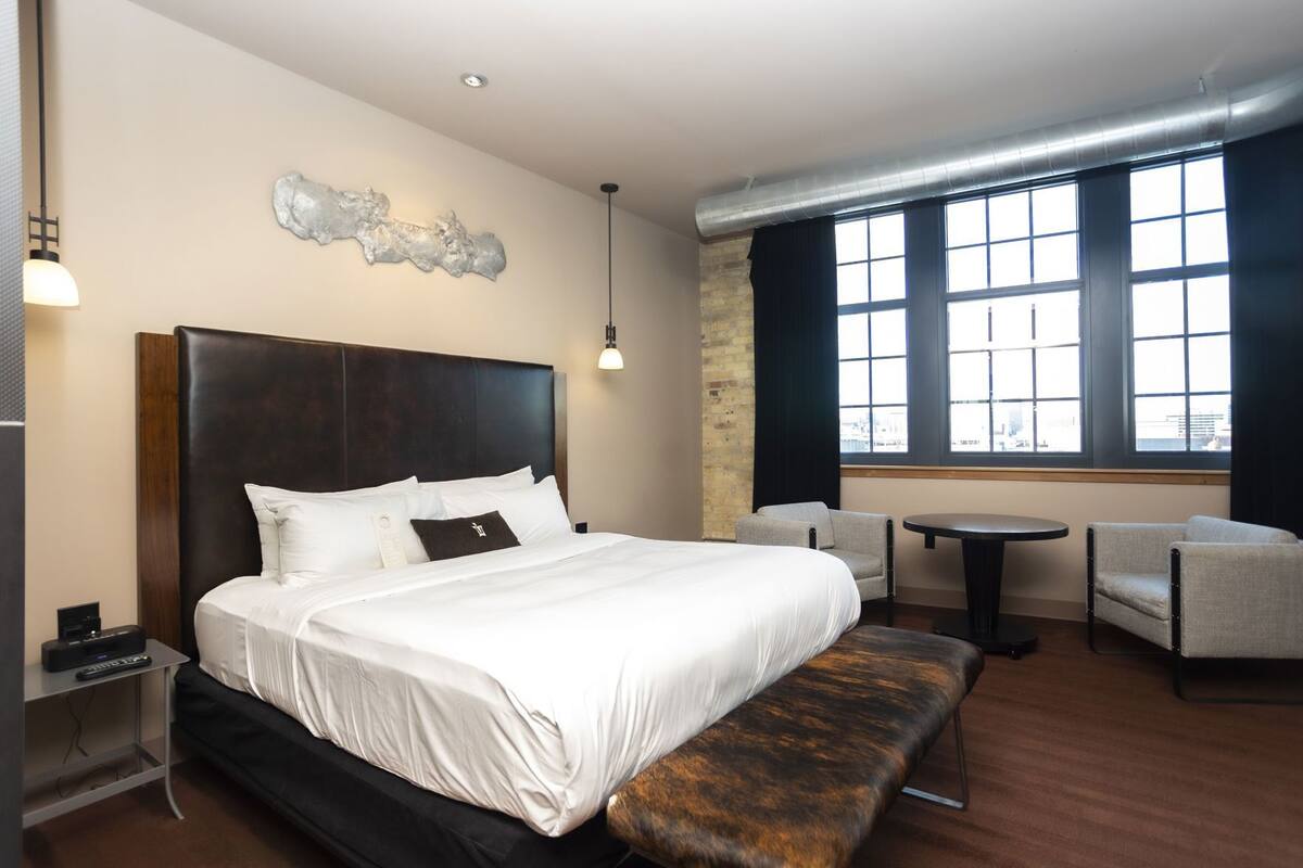 Ein Hotelzimmer mit einem großen Bett, einer Bank, zwei Sesseln, einem kleinen Tisch und Blick auf die Stadt durch große Fenster.
