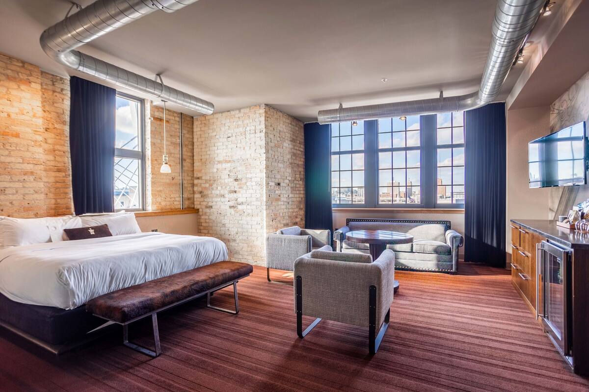 Ein modernes Hotelzimmer mit einem großen Bett, einer Bank, zwei Sesseln, einem kleinen Tisch und einem Fernseher.
