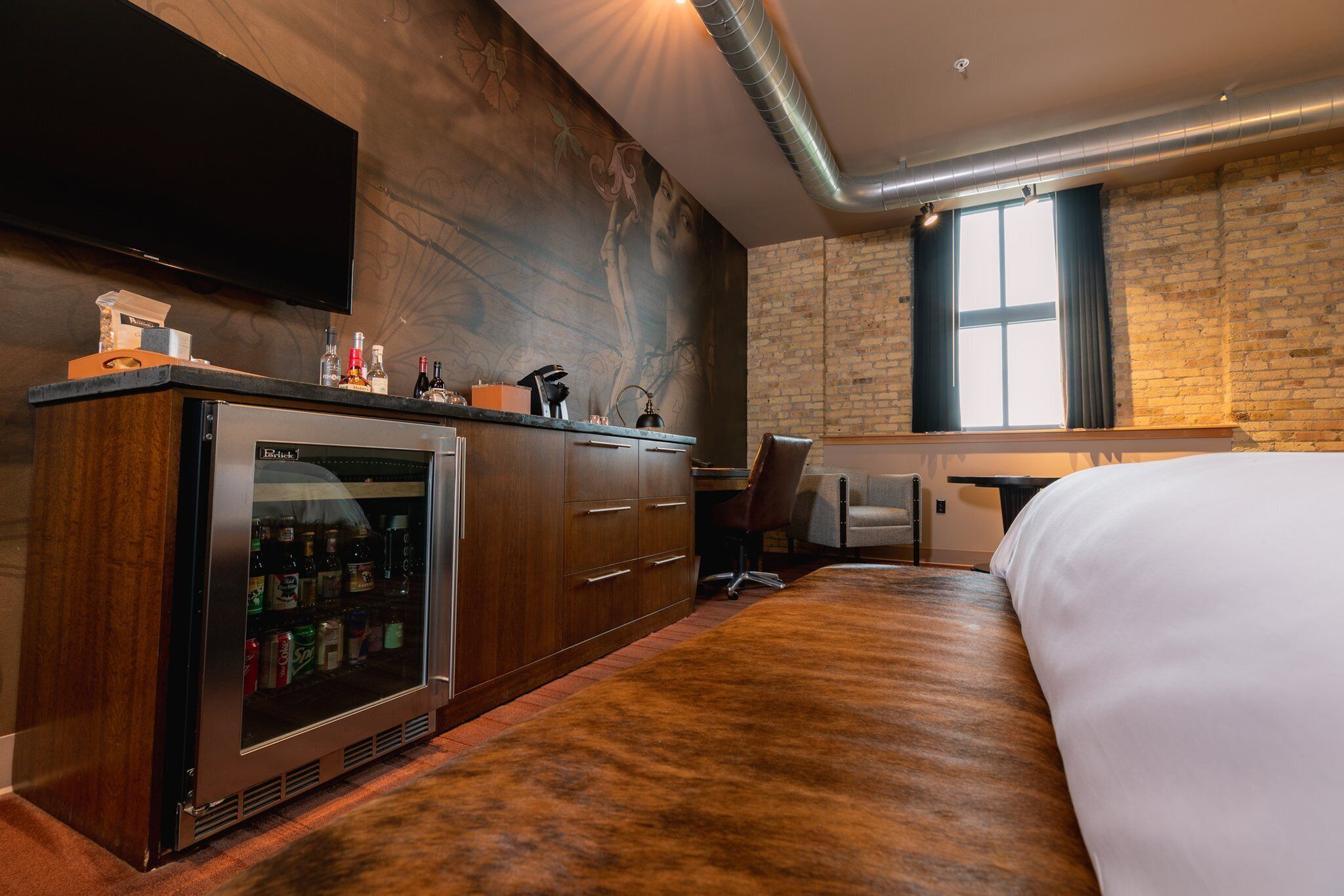 Habitación de hotel con una cama grande, un minibar, un escritorio con una silla, un televisor en la pared y una pared de ladrillo.