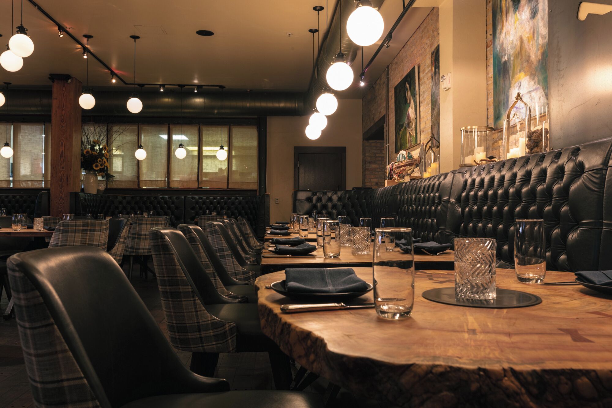 A restaurant with a long wooden table, black leather booth seating, and hanging spherical lights.