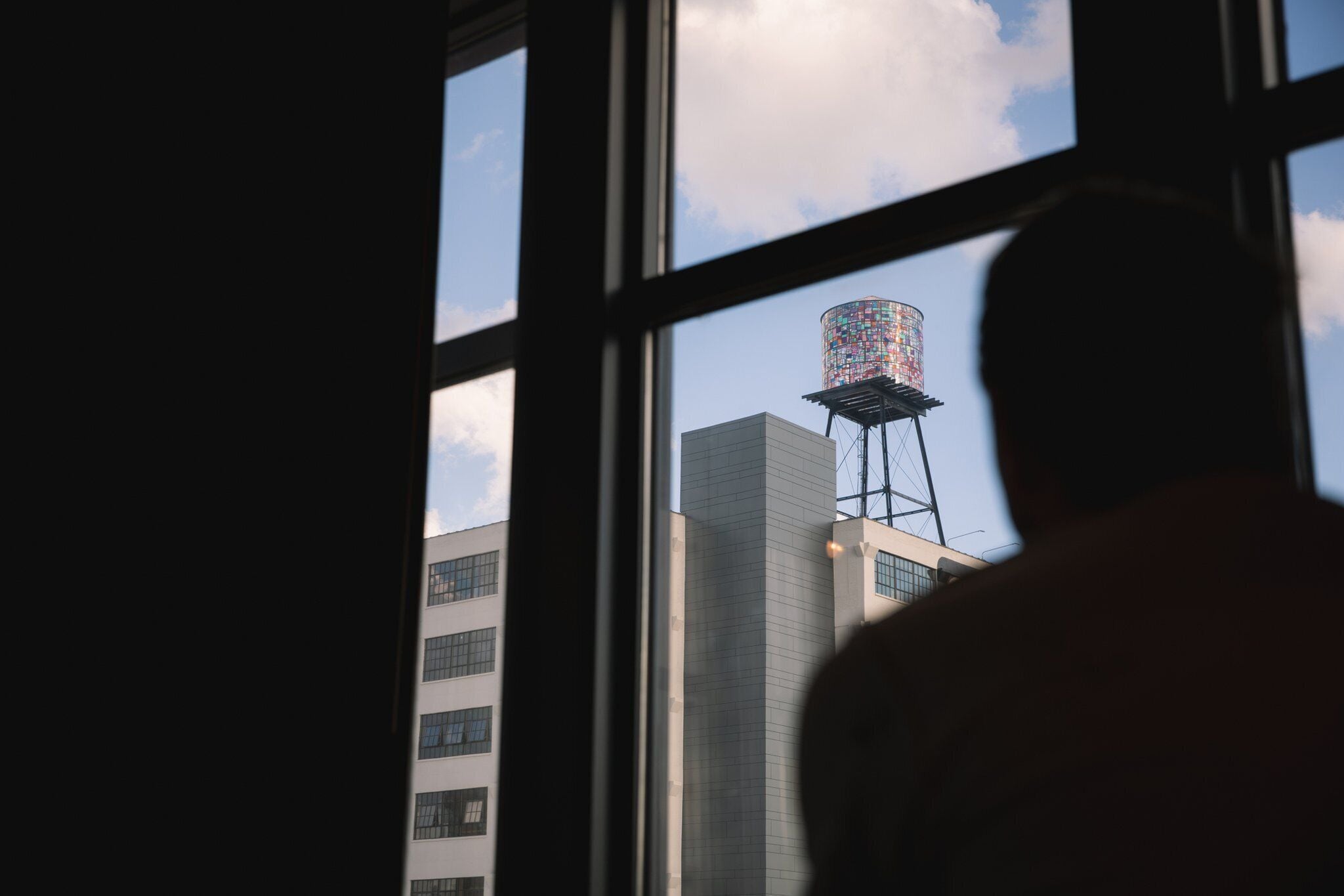 Silueta de una persona de pie frente a una ventana, mirando hacia un edificio alto con una torre de agua colorida.
