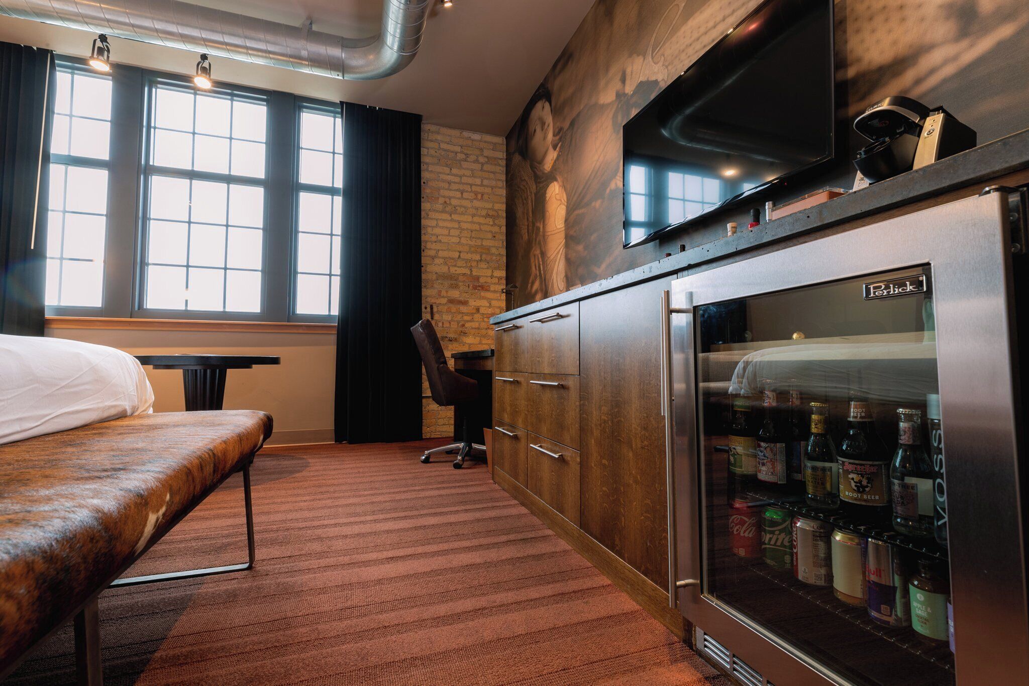 A hotel room with a bed, a minibar, a desk, and a large window.