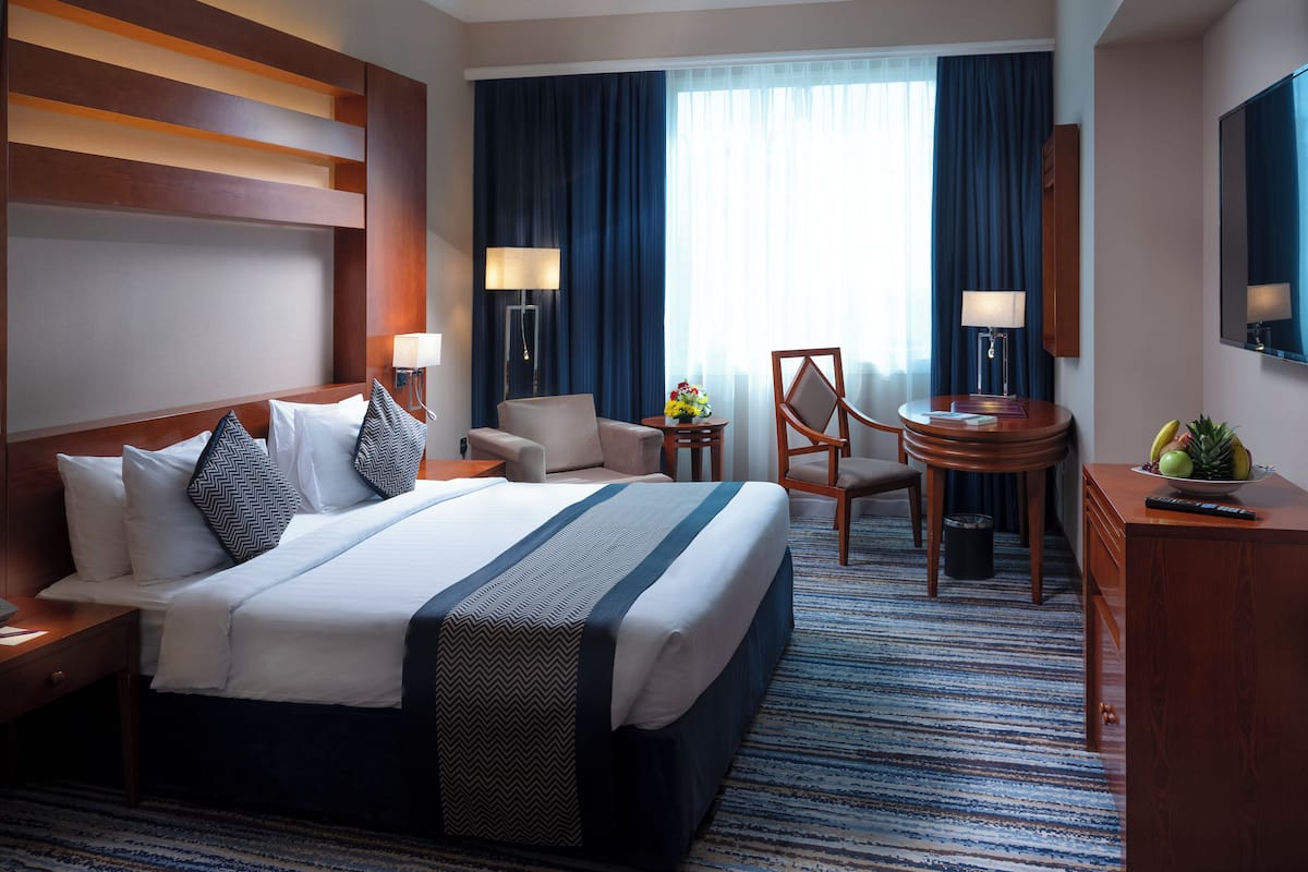 A hotel room with a large bed, a desk, a chair, a small table with a vase of flowers, and a fruit bowl.
