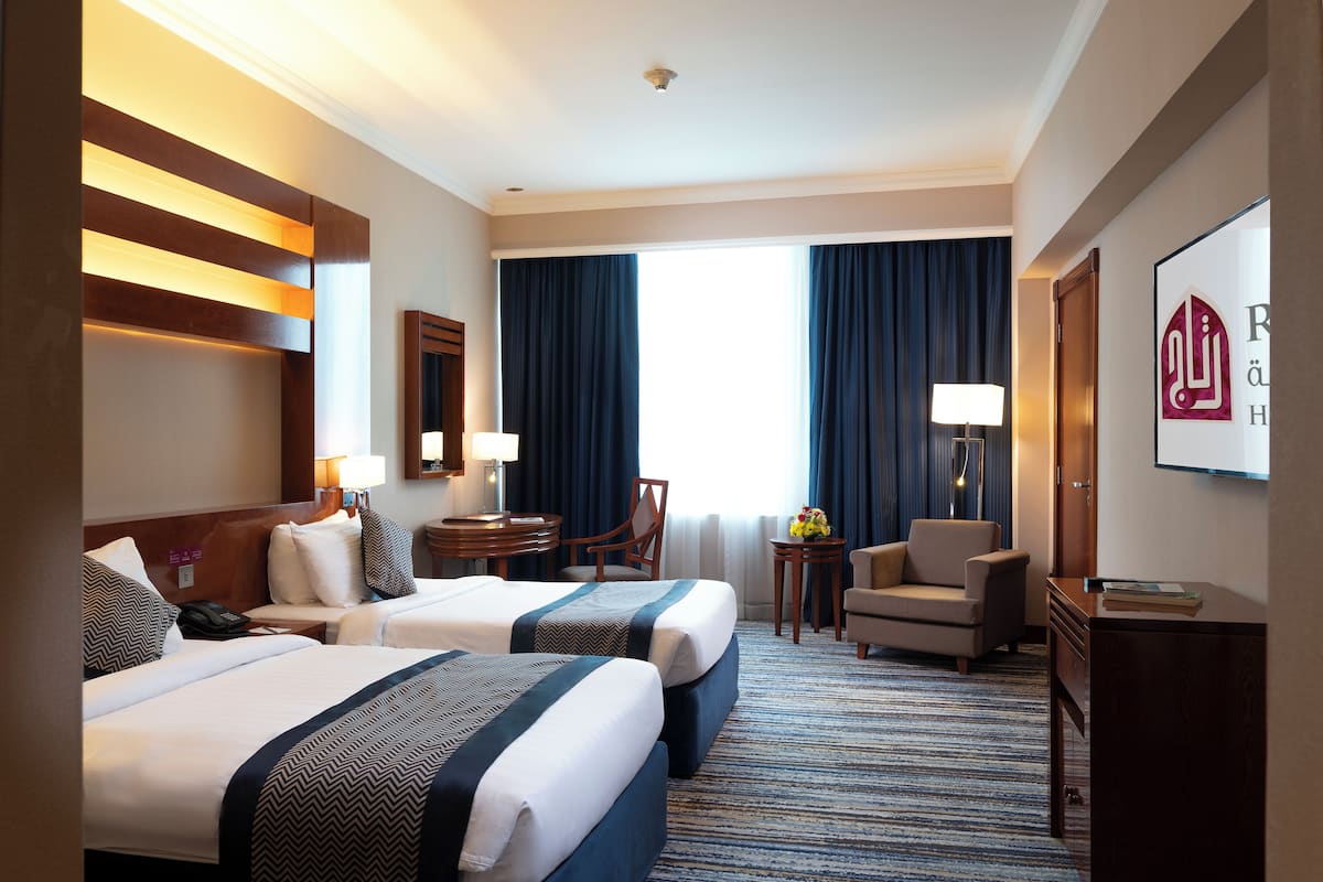 A hotel room with two beds, a desk, a chair, a lamp, and a large window with curtains.