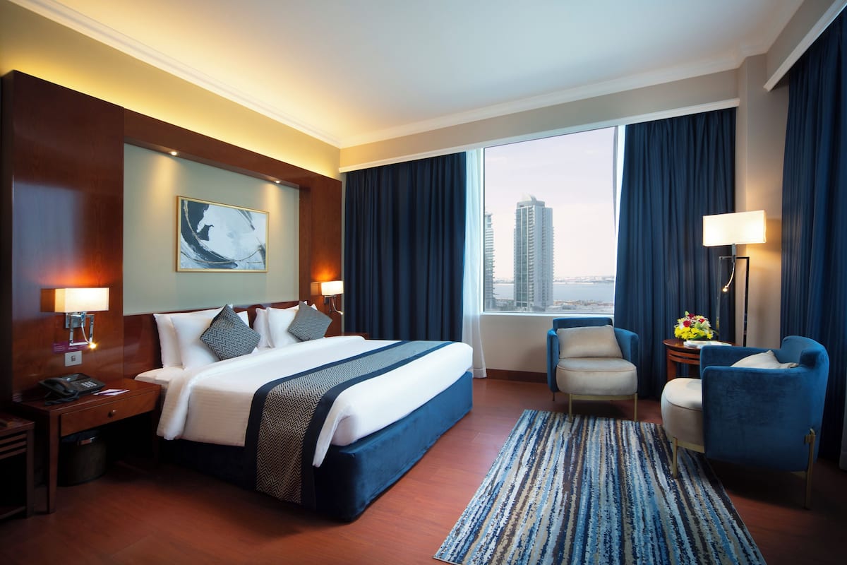 A hotel room with a large bed, a desk with a lamp, a chair, a view of a cityscape, and a window with curtains.