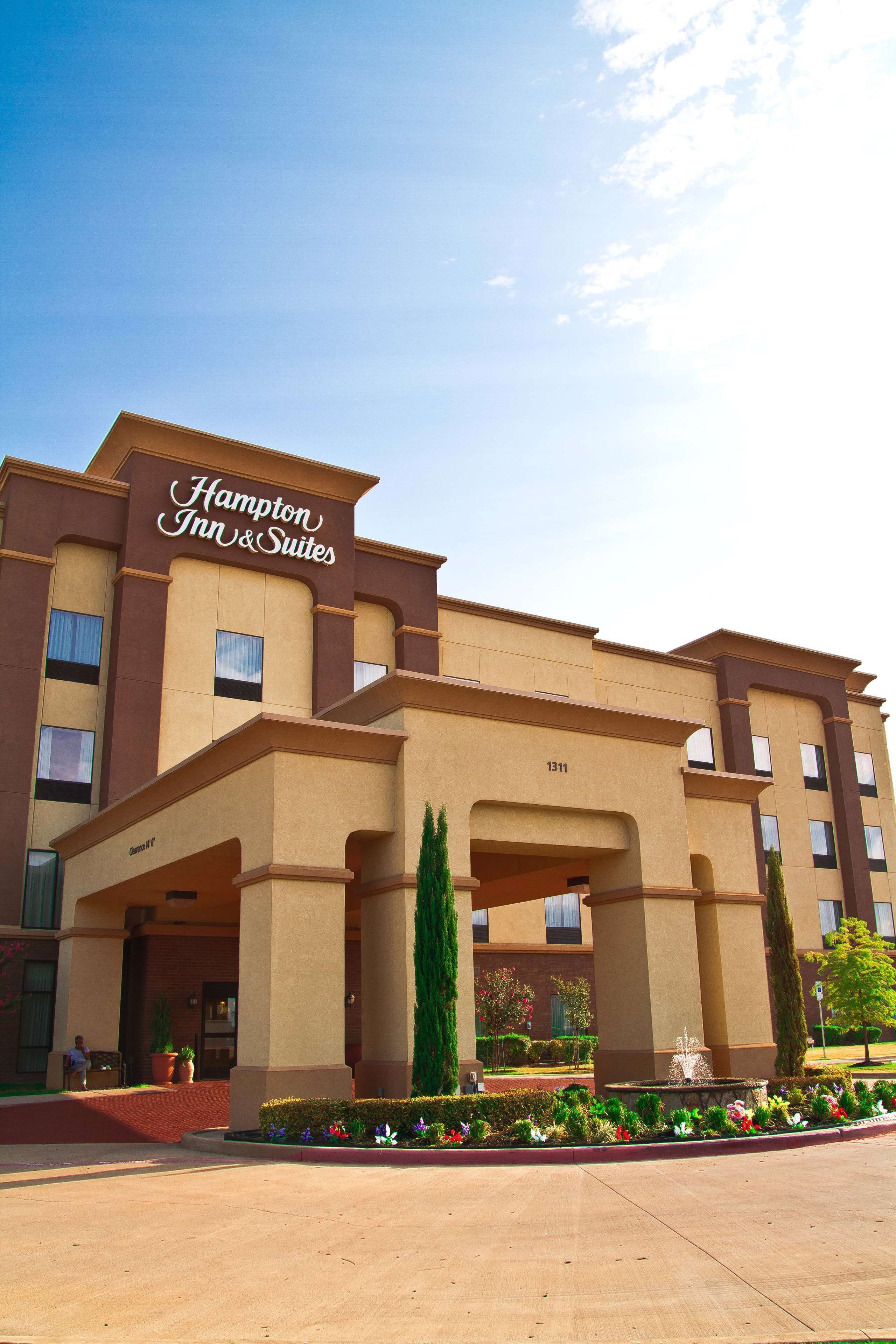 Edificio de Hampton Inn & Suites con una entrada destacada, un jardín ajardinado y un cielo despejado.