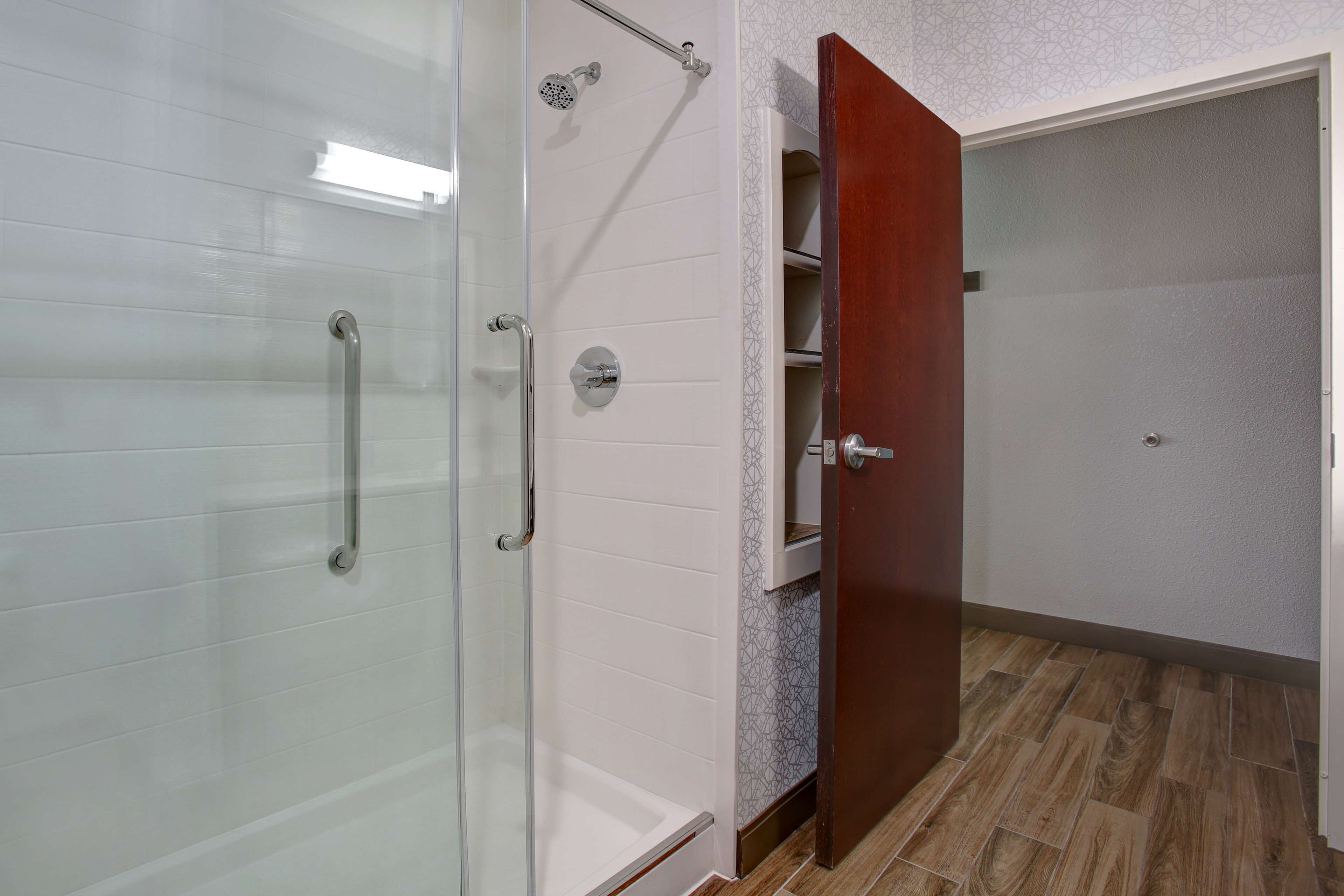 A bathroom with a glass shower enclosure, a white tiled wall, a wooden door, and a patterned floor.