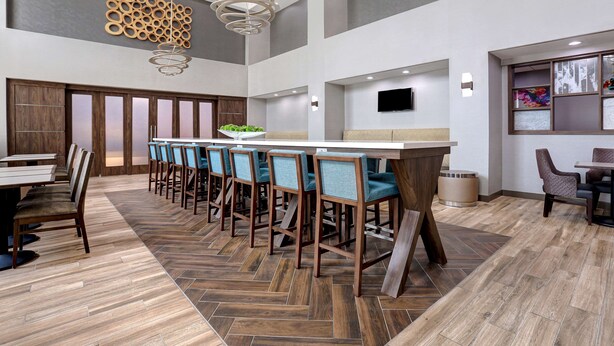 A modern hotel lobby with a long bar area, wooden flooring, and a large wall-mounted TV. - Hampton Inn & Suites Dallas-DeSoto (DeSoto)