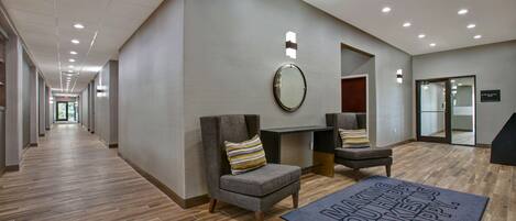 A hotel lobby with a long corridor, seating area with chairs and a small table, a mirror on the wall, and a welcome mat on the floor.