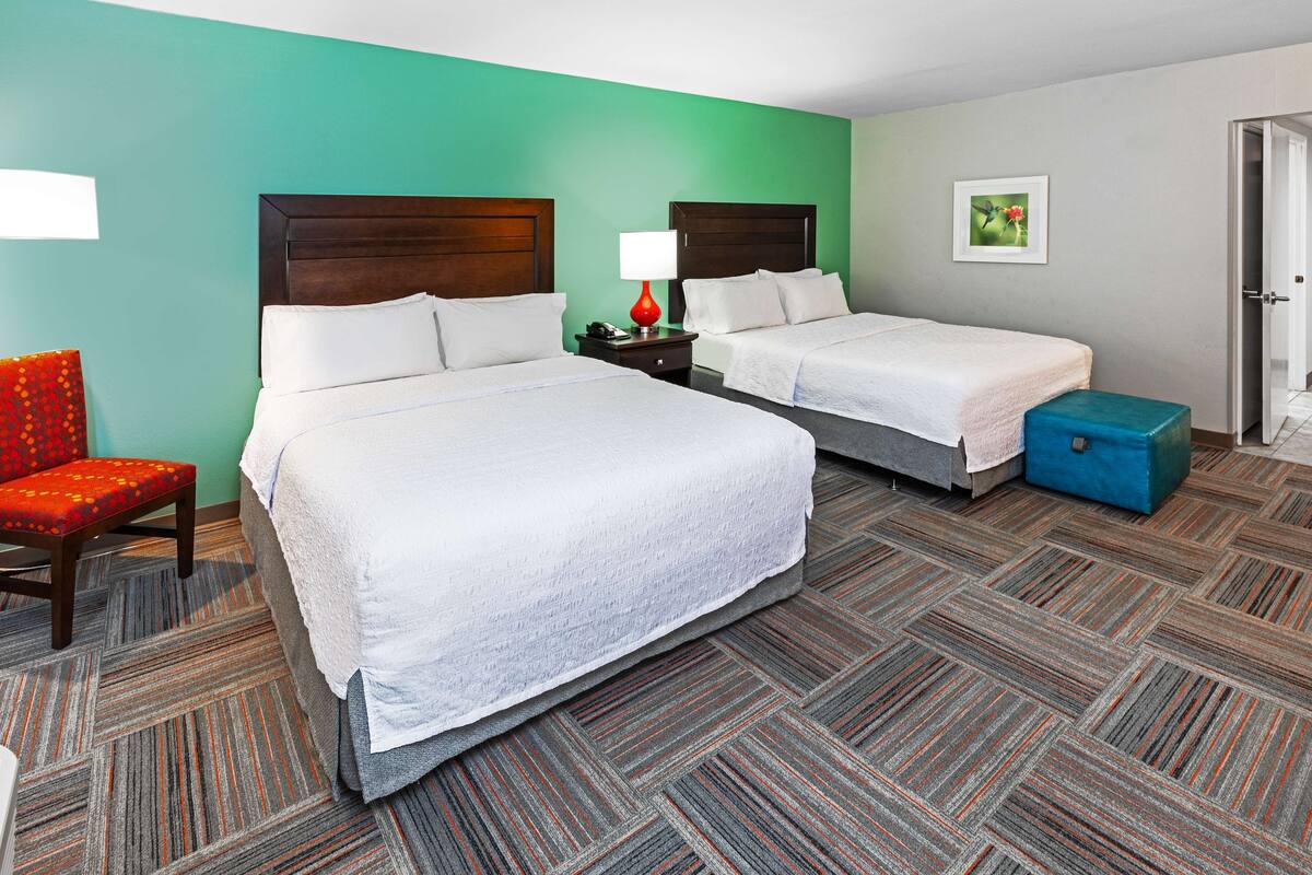 A hotel room with two beds, a red chair, a blue storage box, and a nightstand with a lamp.