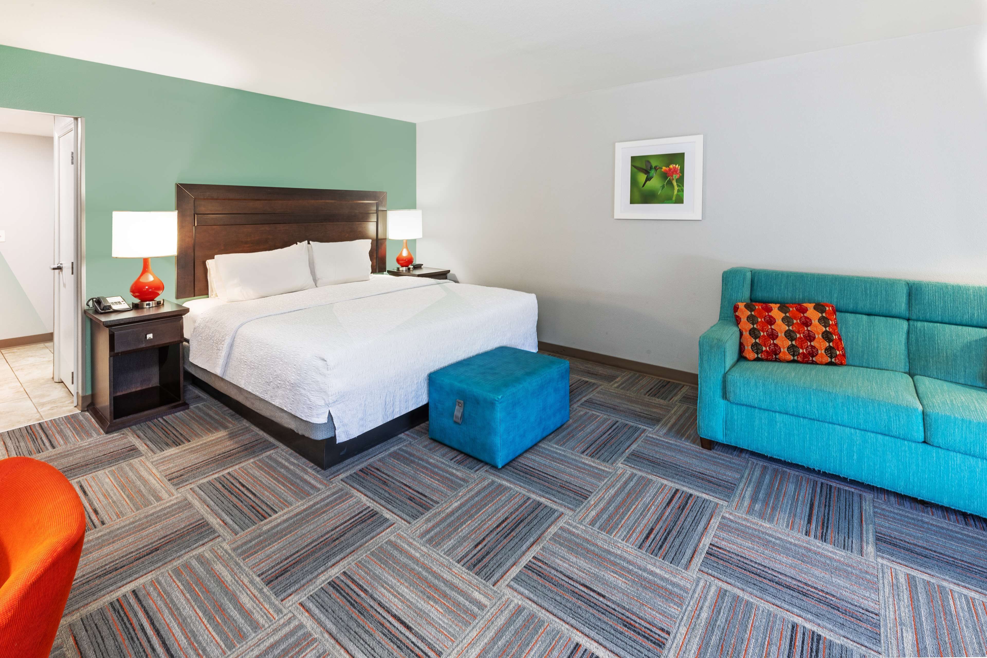 Una habitación de hotel con una cama, un otoman azul, un sofá verde azulado y una mesita de noche con una lámpara.