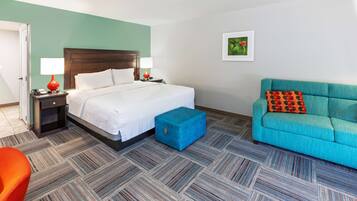 Una habitación de hotel con una cama, un otoman azul, un sofá verde azulado y una mesita de noche con una lámpara.