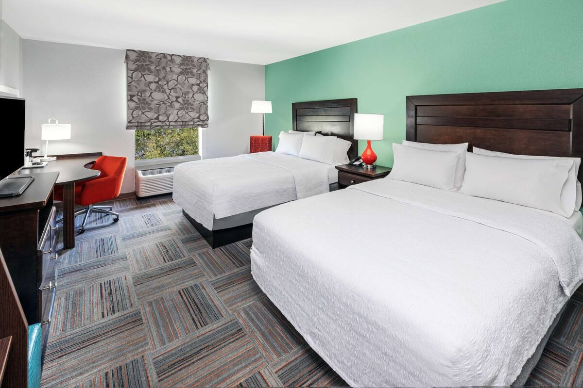 A hotel room with two beds, a desk with a chair, a TV, and a window with a view.