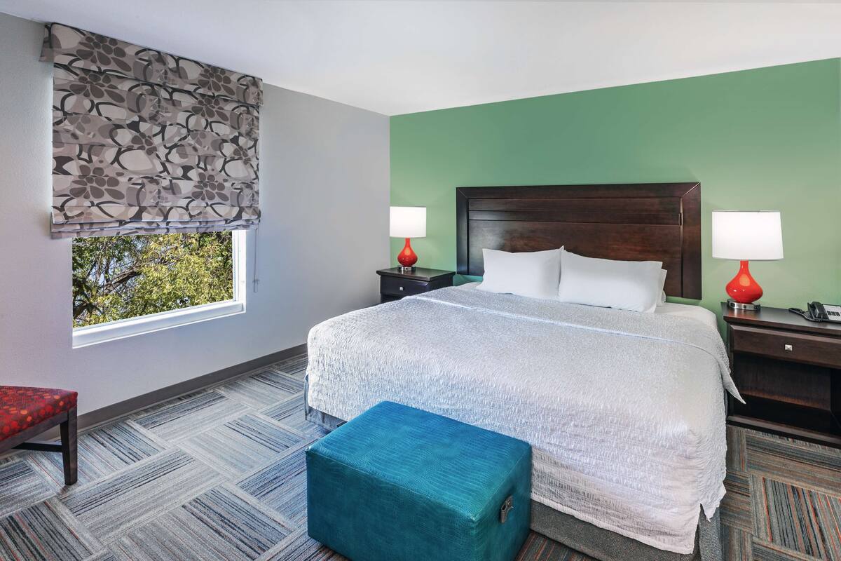 A hotel room with a large bed, two bedside tables with lamps, a bench, a window with a view, and a patterned carpet.