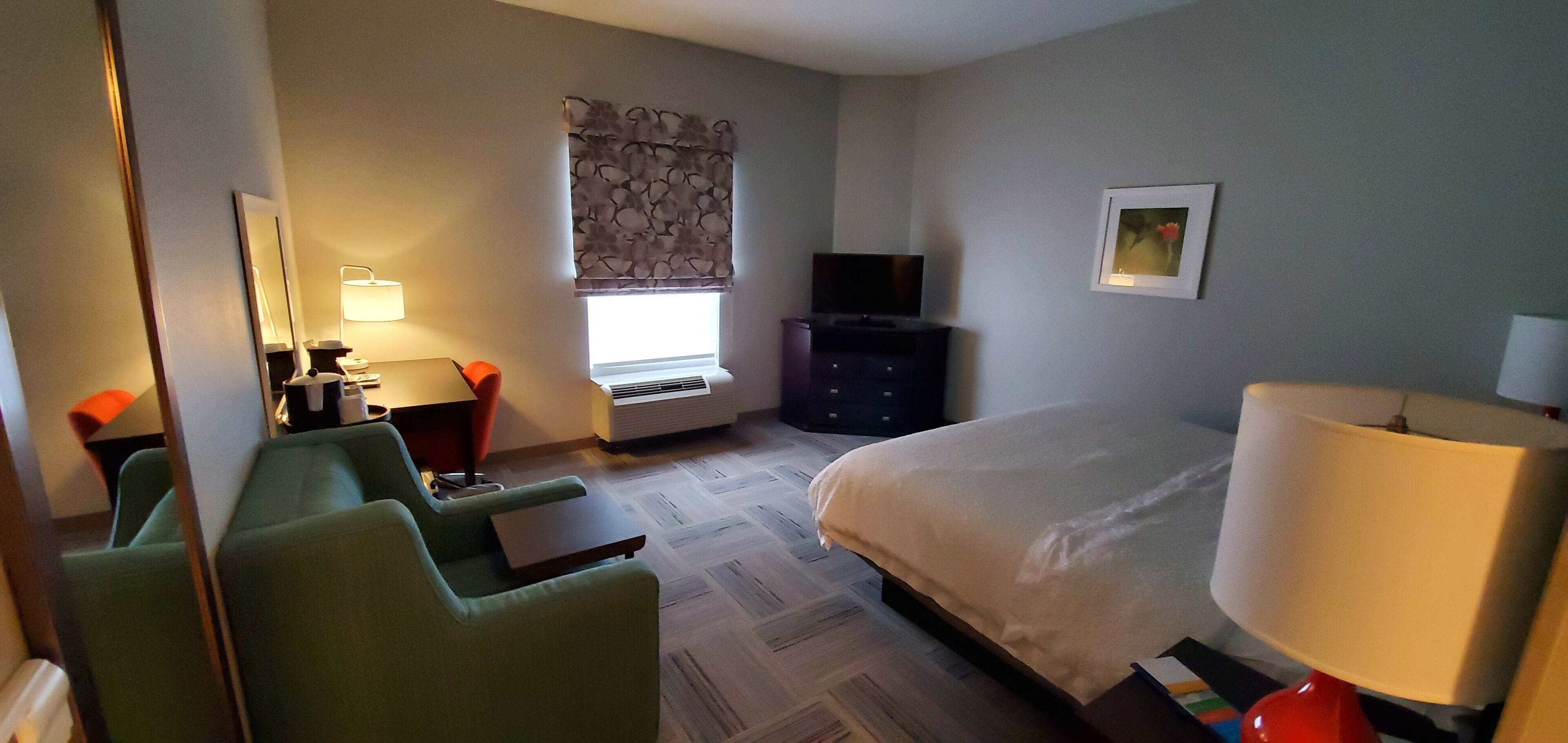 A hotel room with a bed, a desk with a chair, a TV, a window with a view, and a lamp.