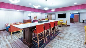 A modern hotel room with a large dining table, multiple chairs, a sofa, and a TV mounted on the wall.