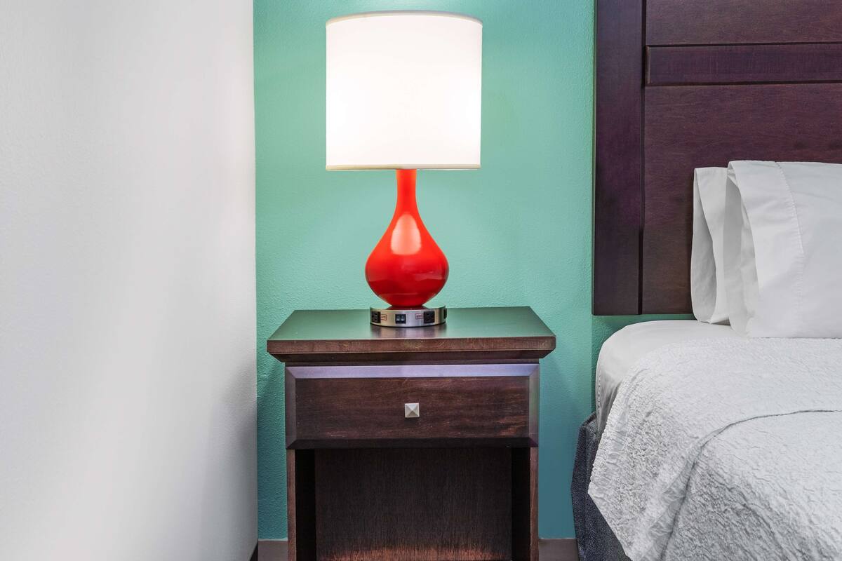A neatly made bed with white linens, a red table lamp on a wooden nightstand, and a teal wall.
