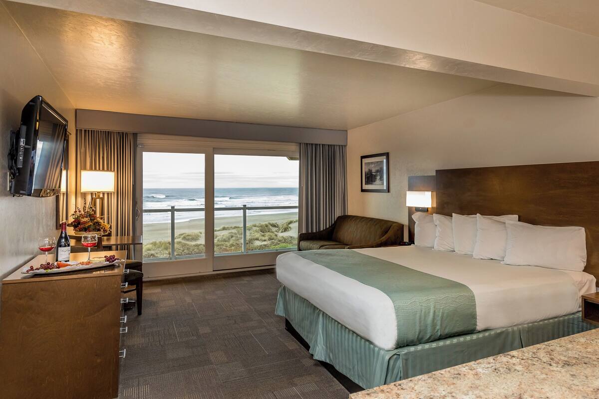 A hotel room with a large bed, a desk with a TV, a chair, a view of the ocean, and a picture on the wall.