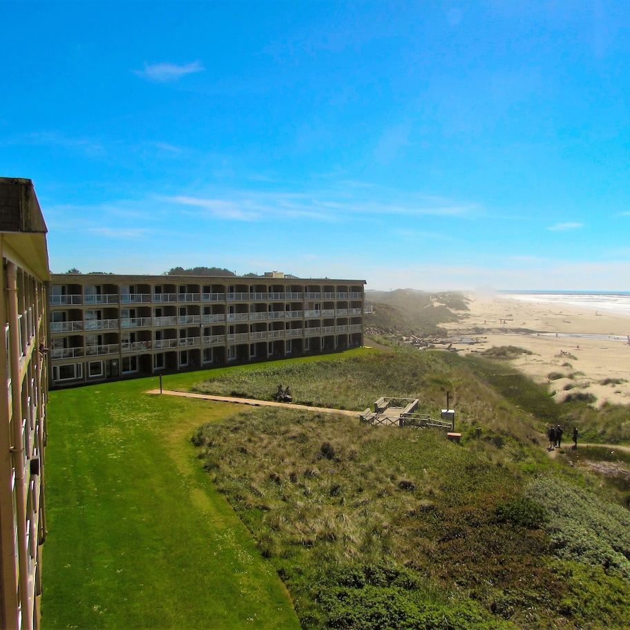 A multi-story building with a flat roof and large windows, overlooking a beach with sand dunes and a clear sky.