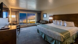 A hotel room with a large bed, a desk, a chair, and a view of the sunset through the window.