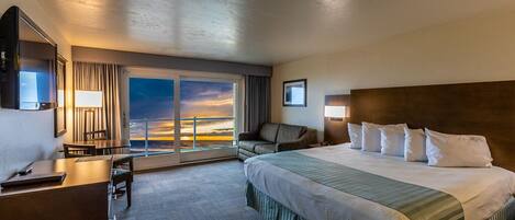 A hotel room with a large bed, a desk, a chair, and a view of the sunset through the window.
